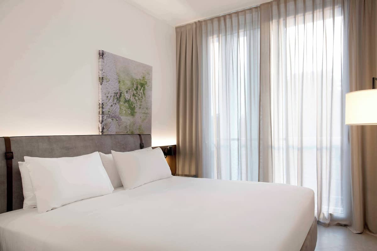 A hotel room with a large bed, a headboard, a wall-mounted artwork, and a window with curtains.
