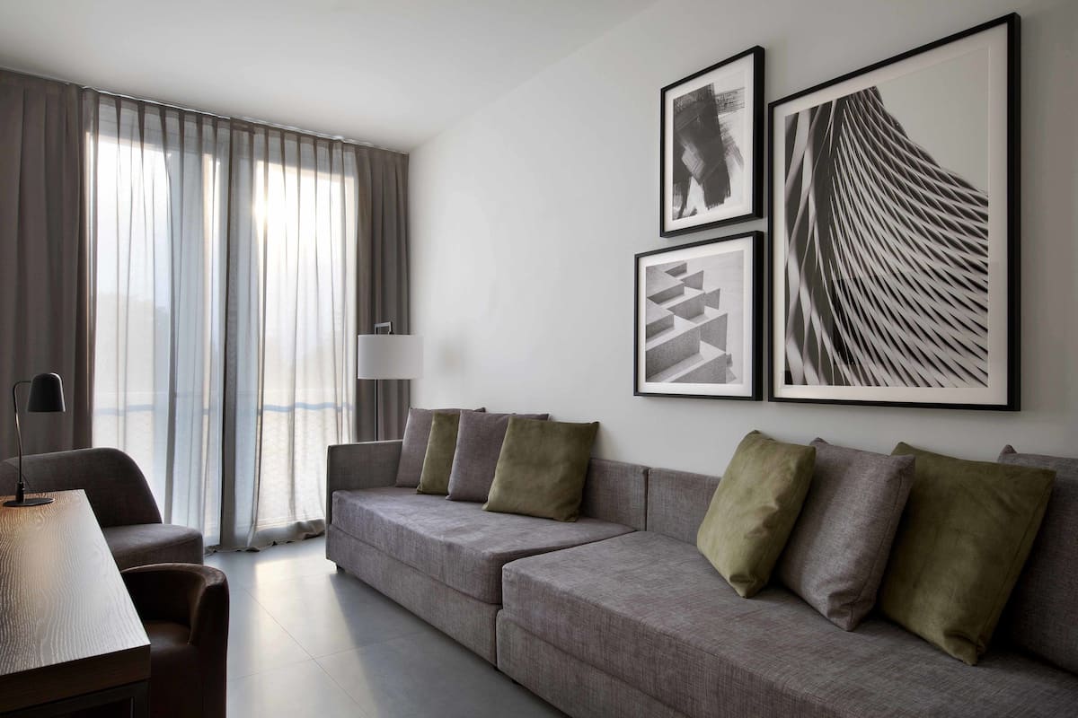 A modern living room with a grey sofa, a small table, and a lamp. There are curtains on the window and three framed artworks on the wall.