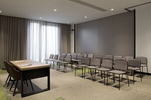 Kokoushuone, jossa on pitkä pöytä ja useita tuoleja, projektorikangas ja suuri ikkuna, jossa on verhot.