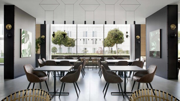 Un espace de salle à manger moderne, doté de luminaires hexagonaux au plafond, de grandes fenêtres donnant sur des arbres et d'une table centrale entourée de chaises.