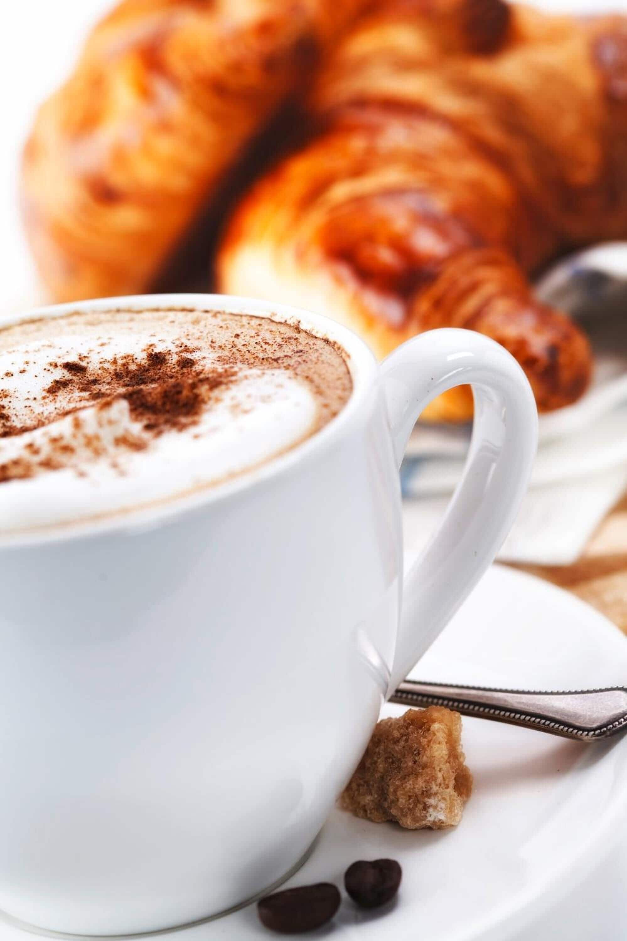 A white cup of coffee with a spoon, a croissant, and a chocolate piece on a white plate.