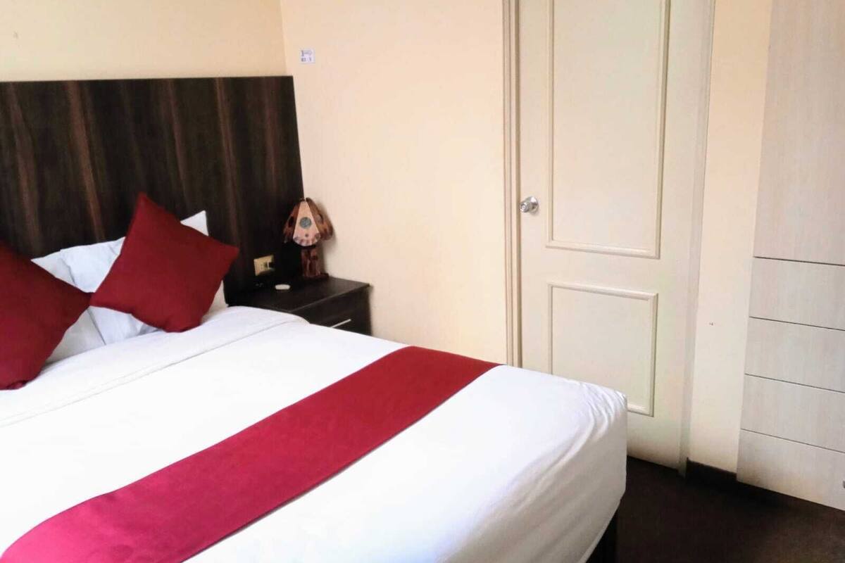 Ein Hotelzimmer mit einem Bett, einem Nachttisch, einem Holzkopfstück, einer Tür und einem an der Wand montierten Lichtschalter.