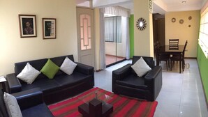 A living room with two black sofas, a glass coffee table, and a red striped rug. There are two framed pictures on the wall, a dining area with a table and chairs, and a green accent wall. - Apart Hotel Incamin (Arequipa)