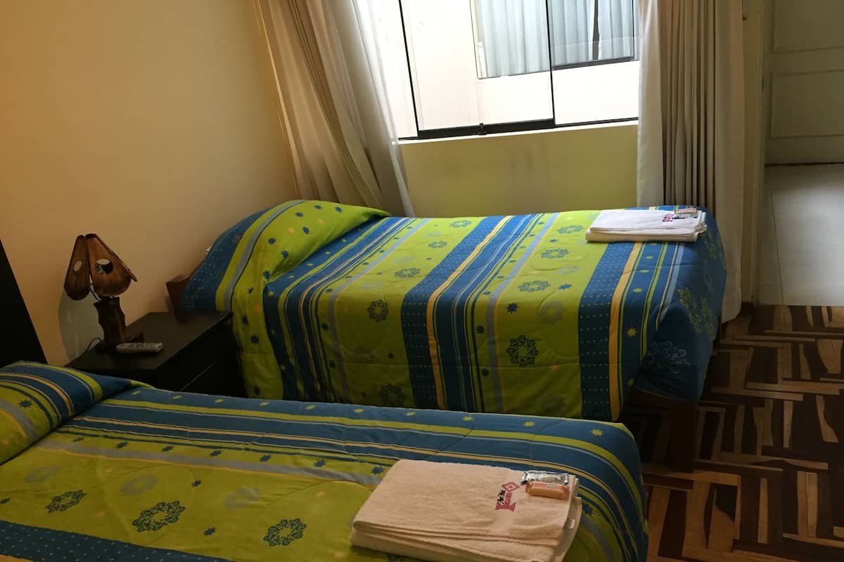 Ein Hotelzimmer mit zwei Betten, jeweils mit einer grün-blauen Streifenoptik, einem Nachttisch mit Lampe und einem Fenster mit Vorhängen.