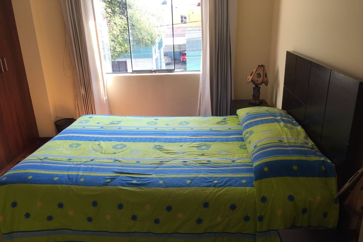 Ein Bett mit gestreifter Bettdecke, ein Nachttisch mit Lampe und ein Fenster mit Vorhängen.