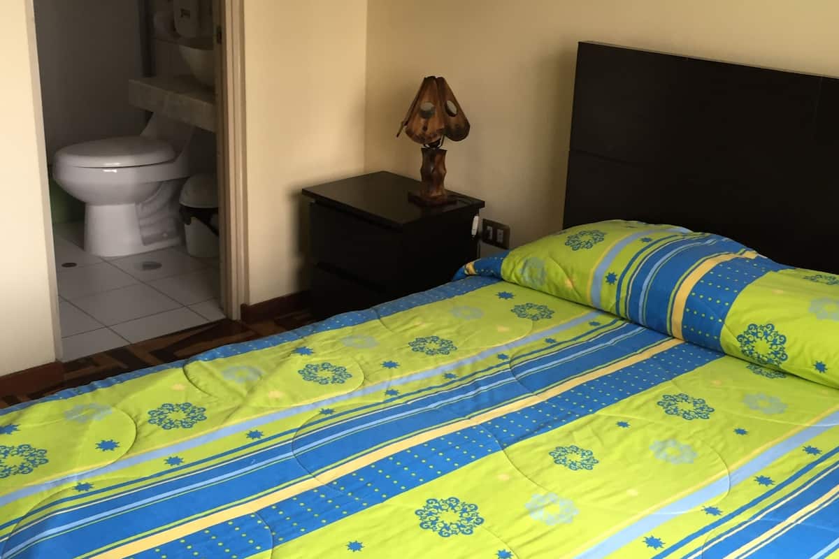 Ein Schlafzimmer mit einem Bett, einem Nachttisch, einer Lampe und einer Toilette, die durch eine offene Tür sichtbar ist.