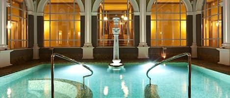 Una amplia piscina interior con una fuente central, rodeada de ventanales de arco y columnas.