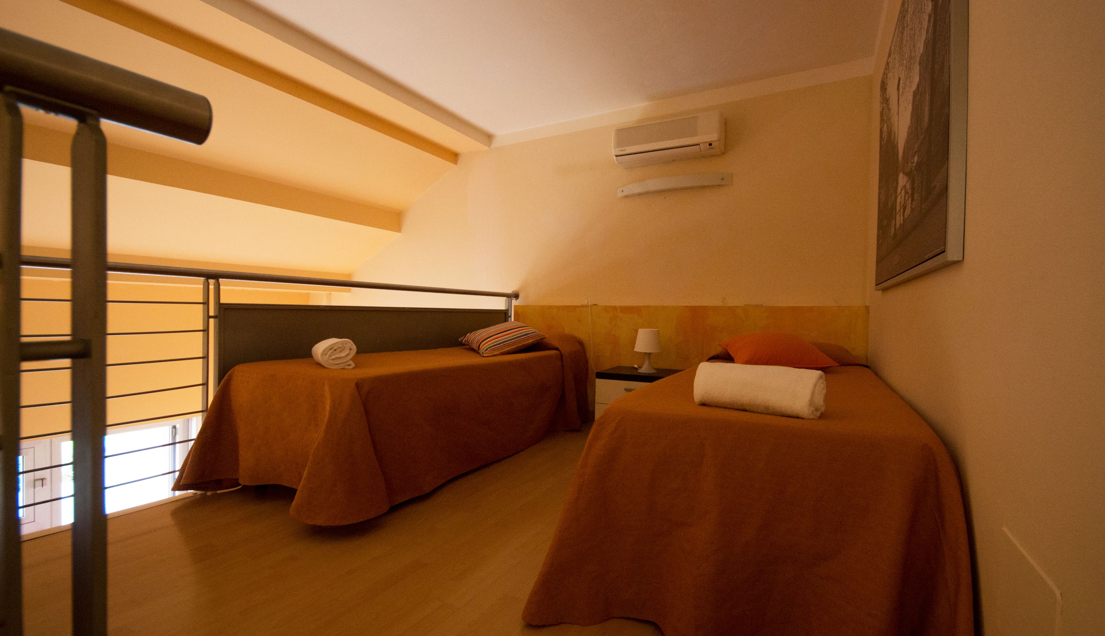 A hotel room with two beds, each with a pillow and a rolled towel. There is a nightstand with a lamp and a wall-mounted air conditioner.
