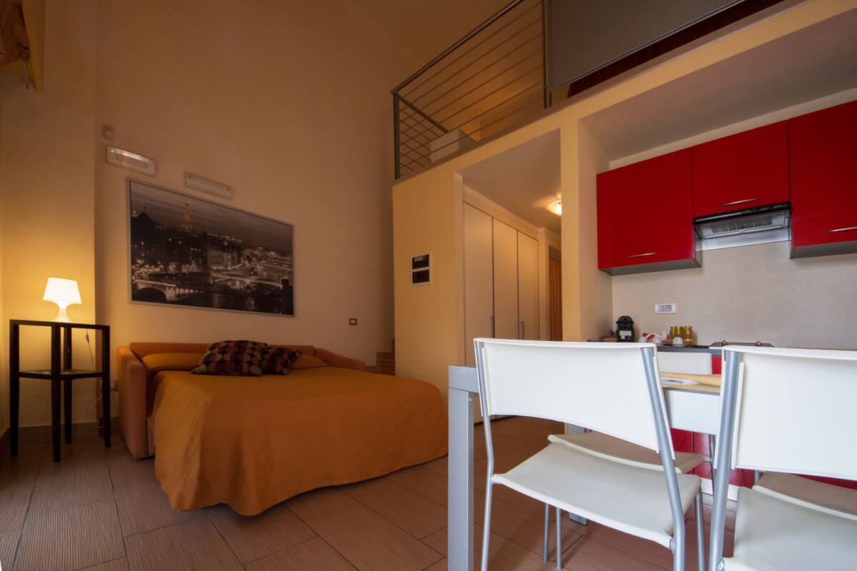 A compact hotel room with a bed, a desk, a chair, a lamp, and a red kitchen area.