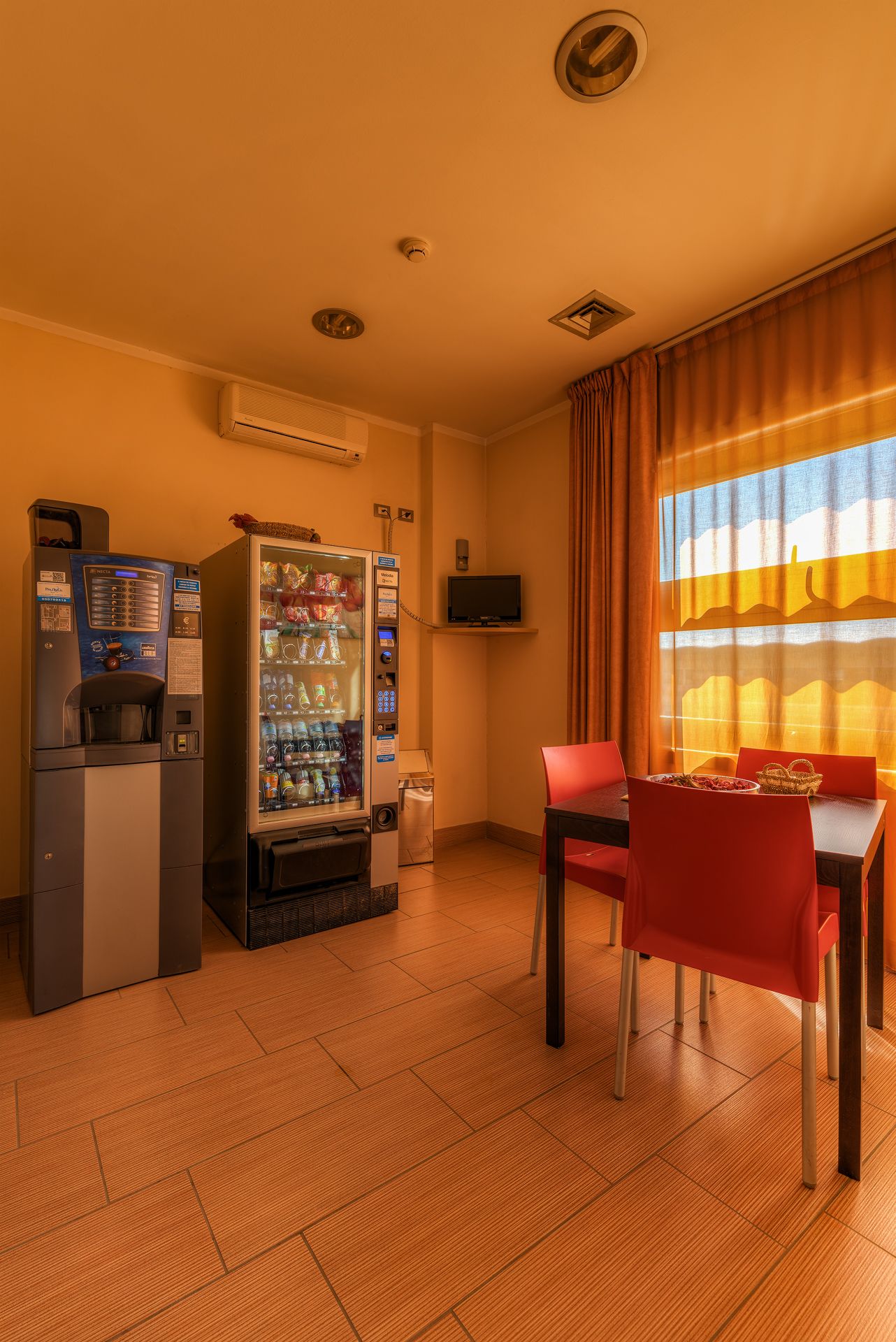 Ein Hotelzimmer mit Minibar, Getränkeautomat, Fernseher und Essecke mit Tisch und Stühlen.