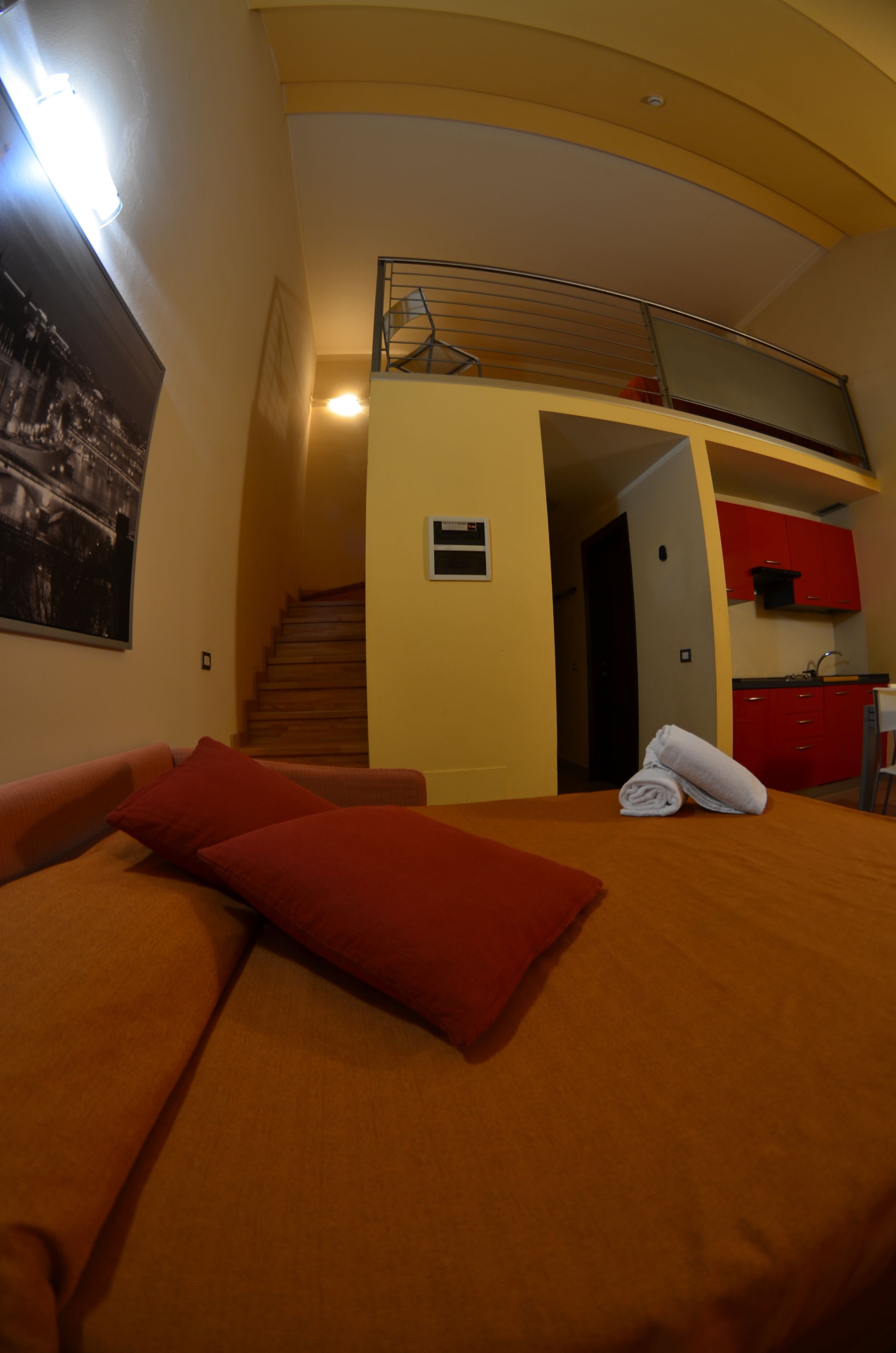 Una camera d'albergo con un letto, un cuscino rosso, un asciugamano arrotolato, una scala, un angolo cottura e un quadro appeso al muro.