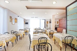 A dining area with multiple tables and chairs, arranged in rows, with a kitchen visible in the background. - Hotel Brezza (Rimini)
