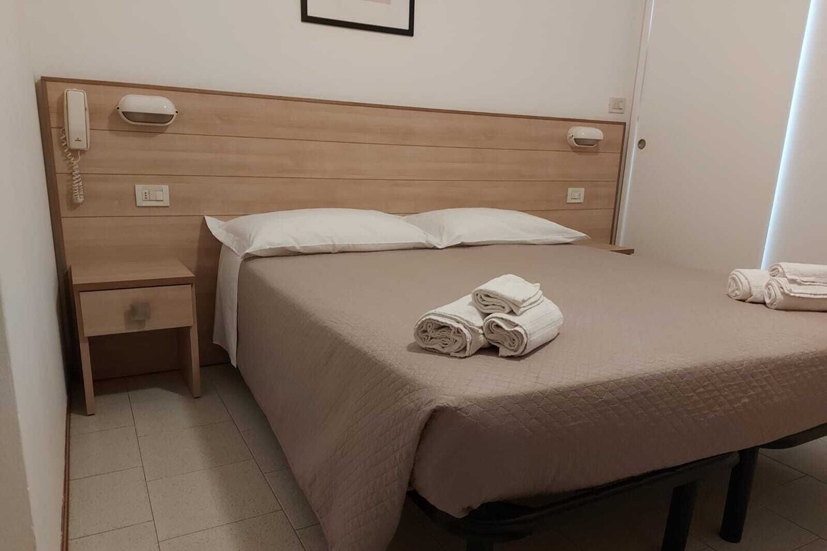 A neatly made bed with a beige bedspread and pillows, a wooden nightstand with a telephone, and a folded towel on the bed.