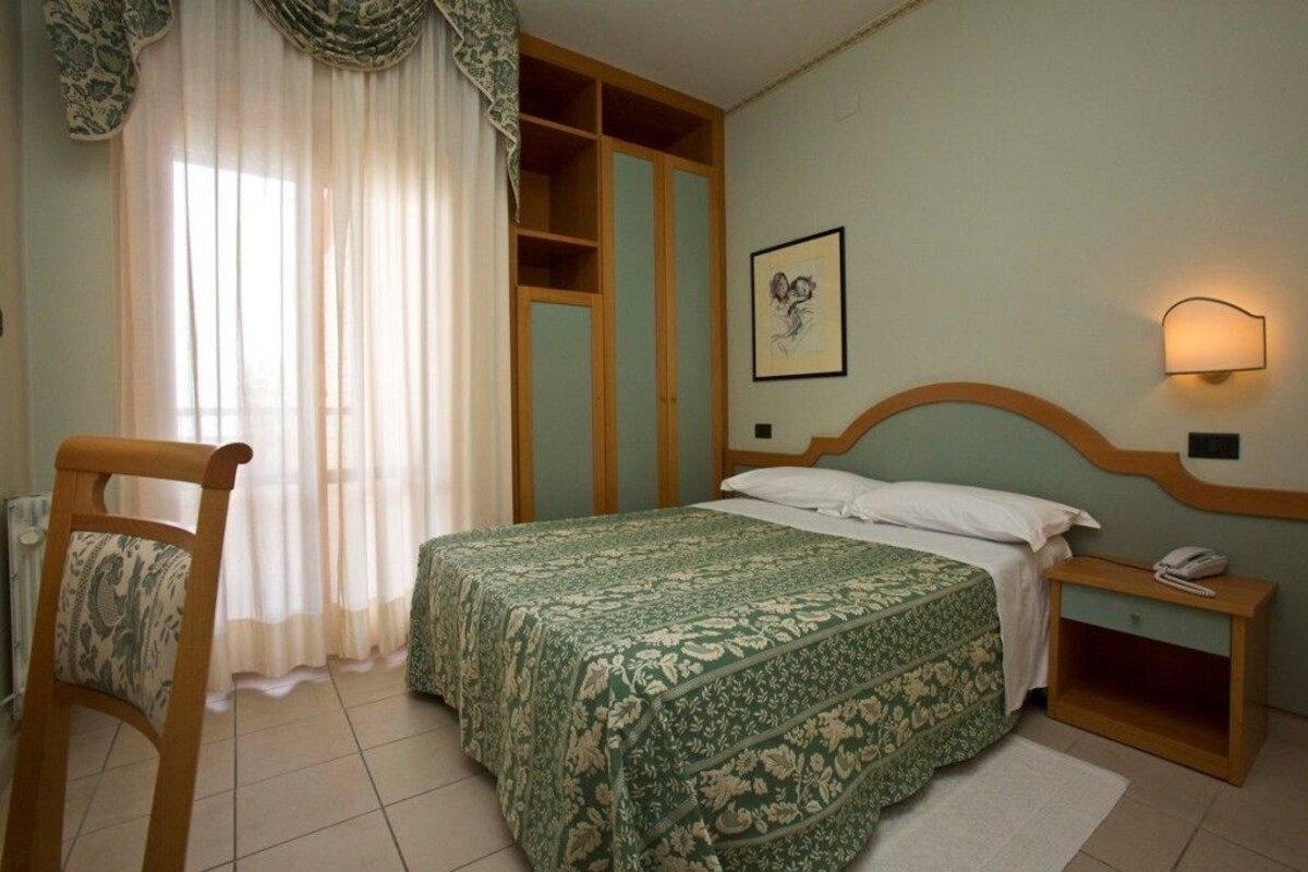 A bedroom with a bed, a chair, a bedside table, a nightstand, a lamp, and a window with curtains.