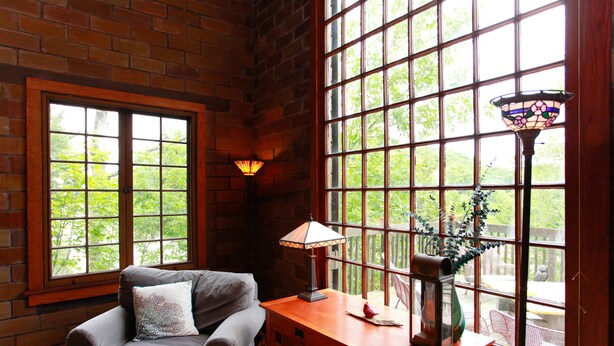 A cozy room with a brick wall, a wooden table, a cushioned chair, and large windows with a view of greenery. - River's Bend (Saugatuck)