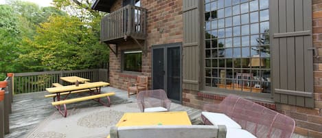 An outdoor patio with picnic tables and wicker chairs, a brick building with large windows, and a wooden deck.