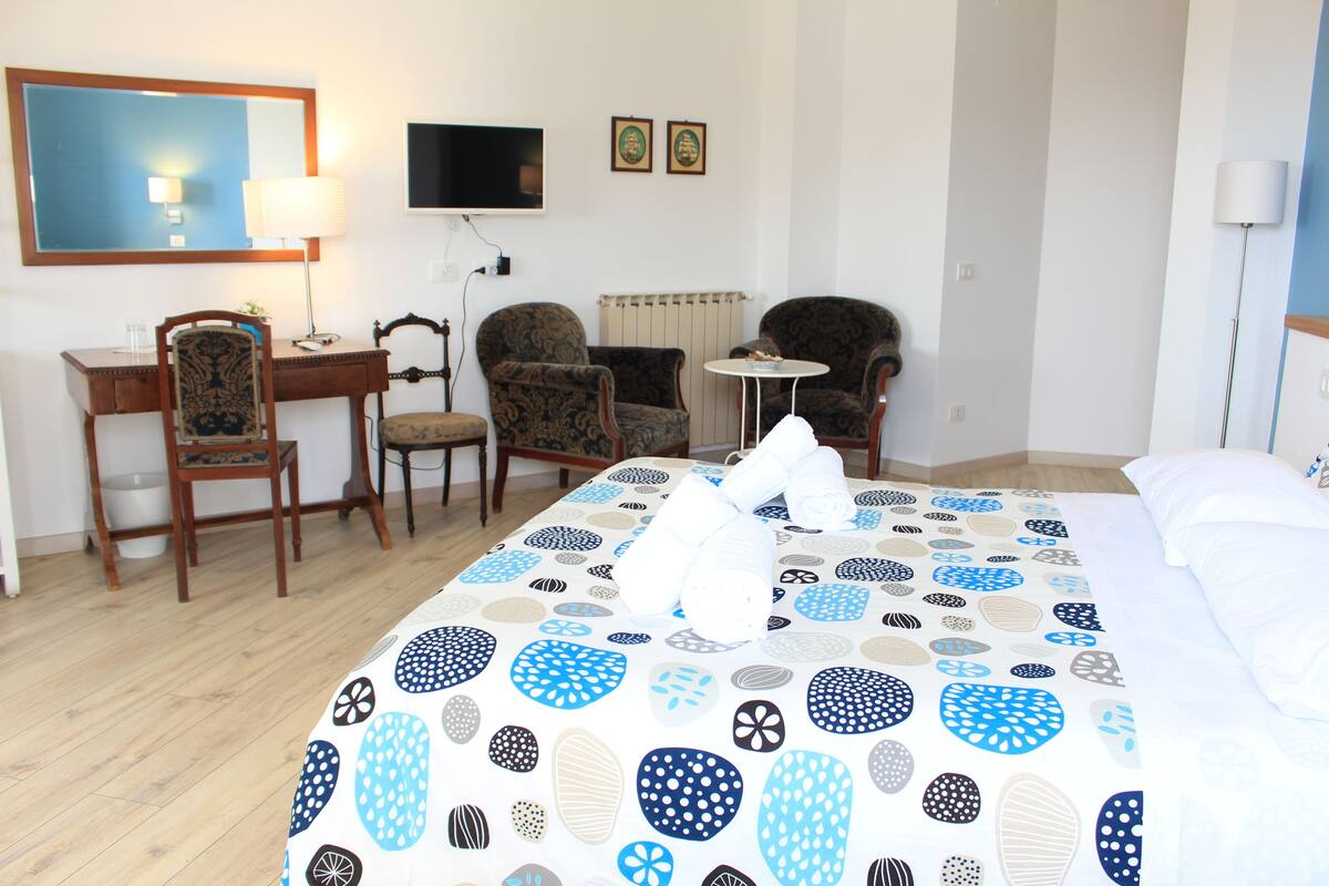 A hotel room with a bed, two armchairs, a desk, a small table, a TV, and a mirror.