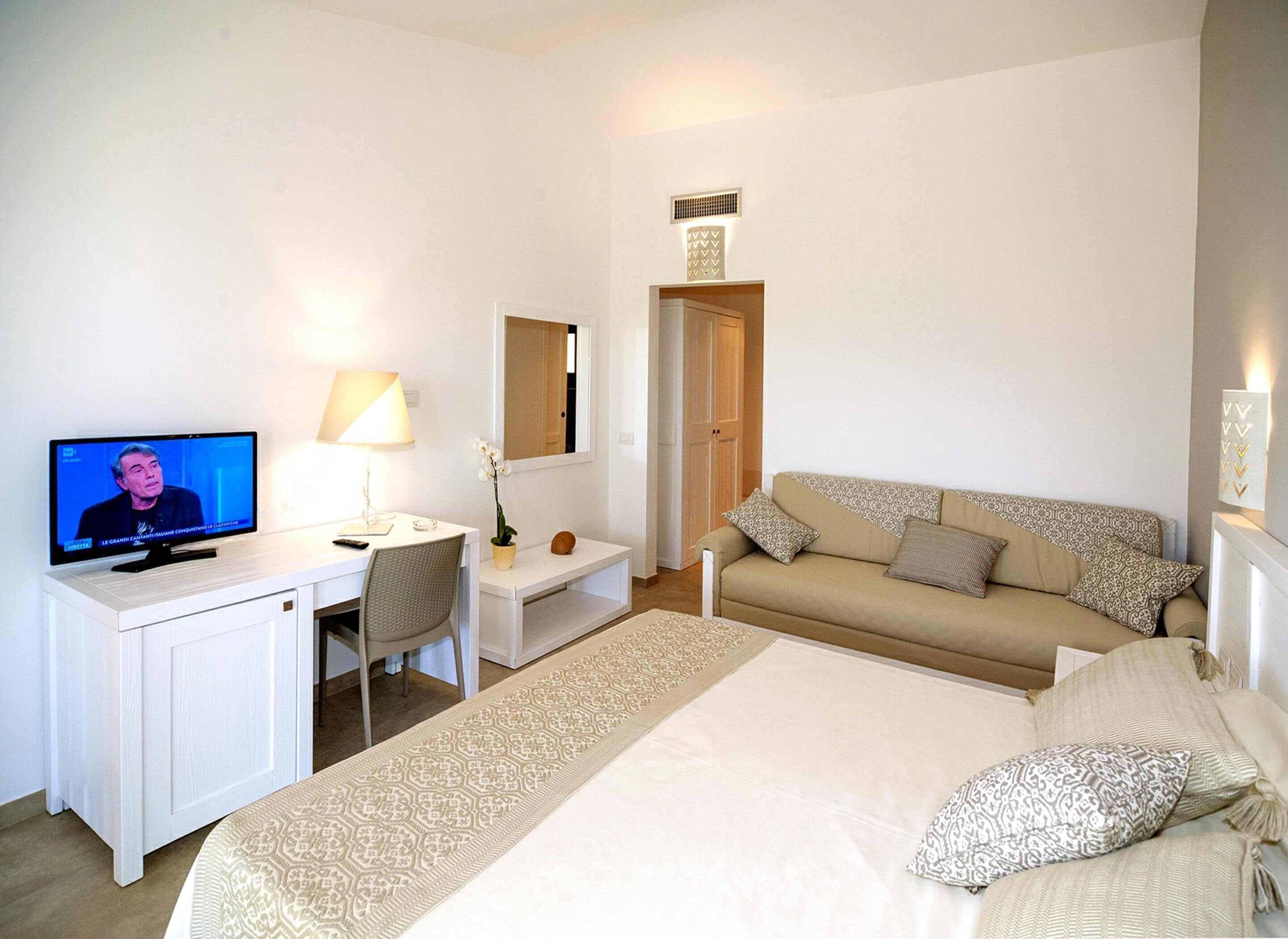 Una habitación de hotel con cama, escritorio con computadora, silla, sofá y televisor mostrando un noticiero.
