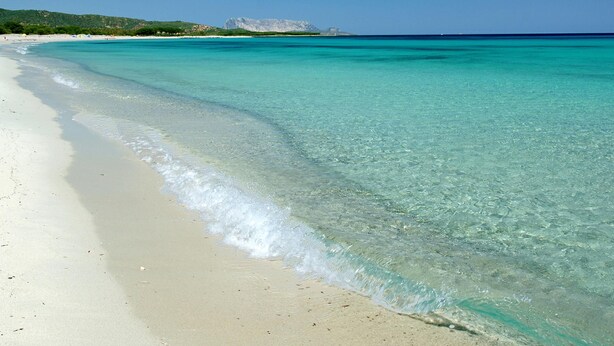 A beach with clear turquoise water, white sand, and a distant rocky outcrop. - Stella Marina Camere sul mare (Budoni)