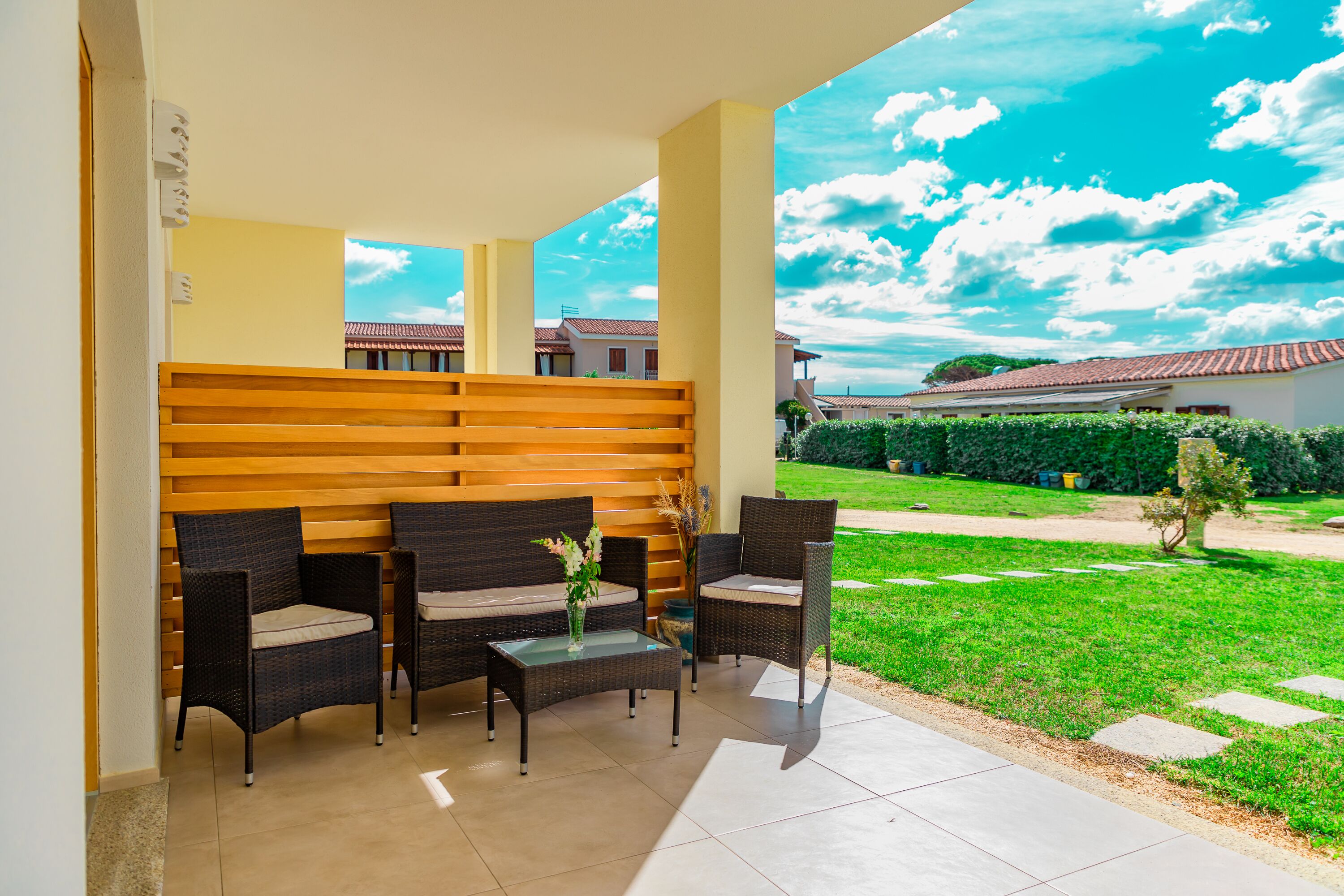 A covered outdoor seating area with wicker furniture and a glass table, overlooking a well-maintained lawn and buildings in the background.