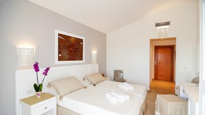 A hotel room with a large bed, two bedside tables, a nightstand, a chair, a small table, and a door. - Stella Marina Camere sul mare (Budoni)