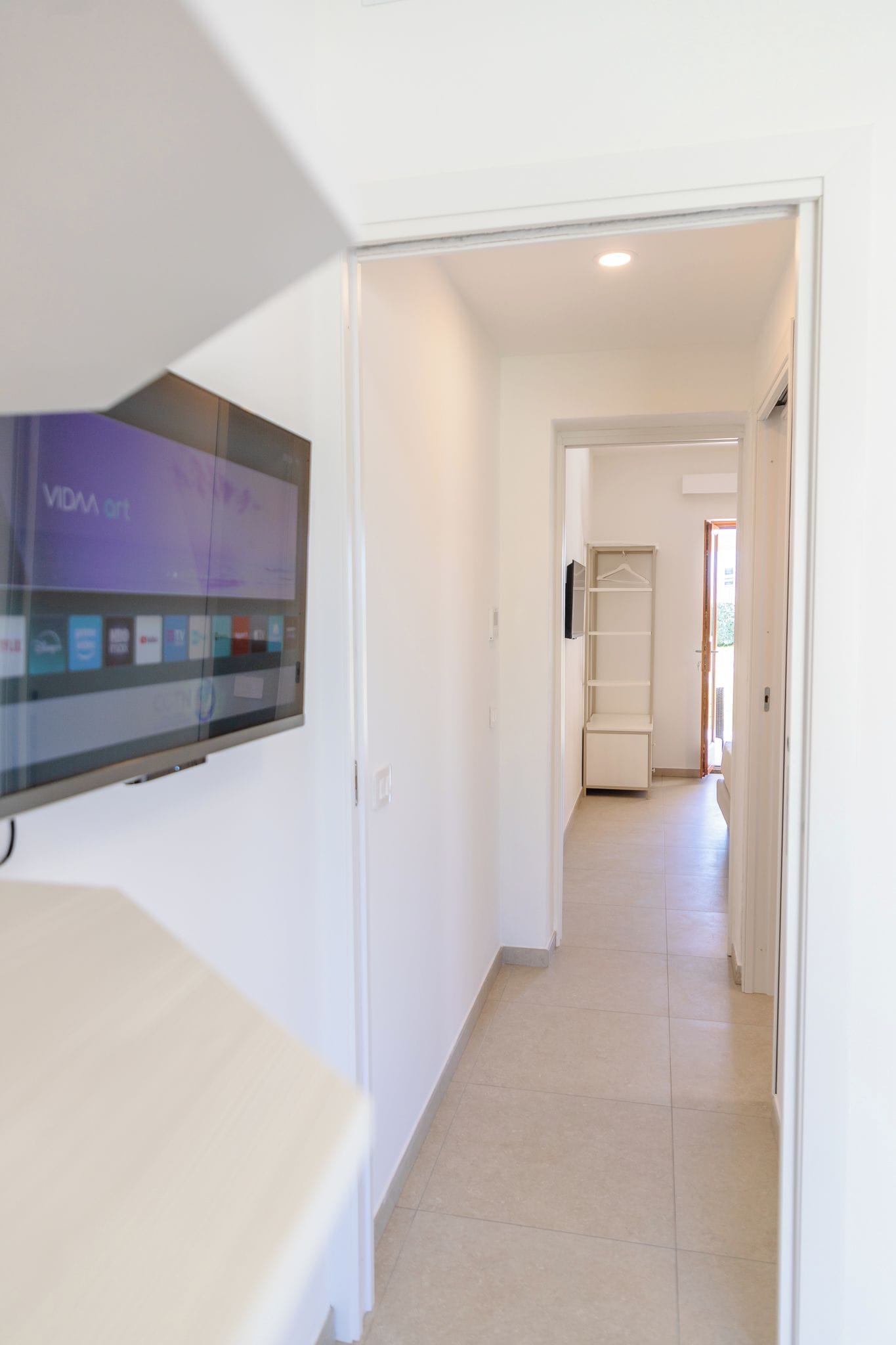 A hallway with a flat-screen TV mounted on the wall, a white door, and a tiled floor.