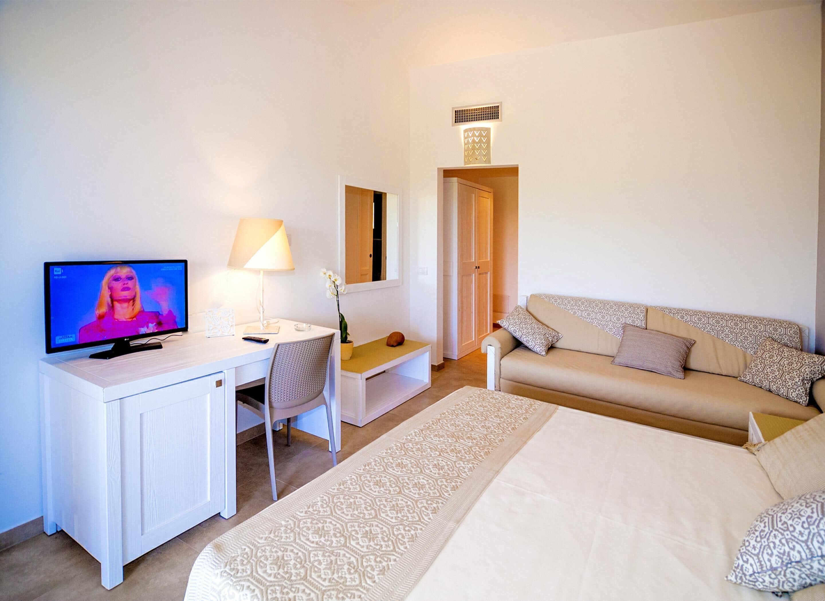 A hotel room with a bed, a desk with a TV, a chair, a sofa, and a lamp.