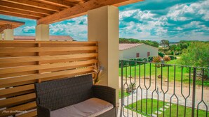 A wooden balcony with a wicker sofa and a view of a green landscape and a red-roofed building. - Stella Marina Camere sul mare (Budoni)