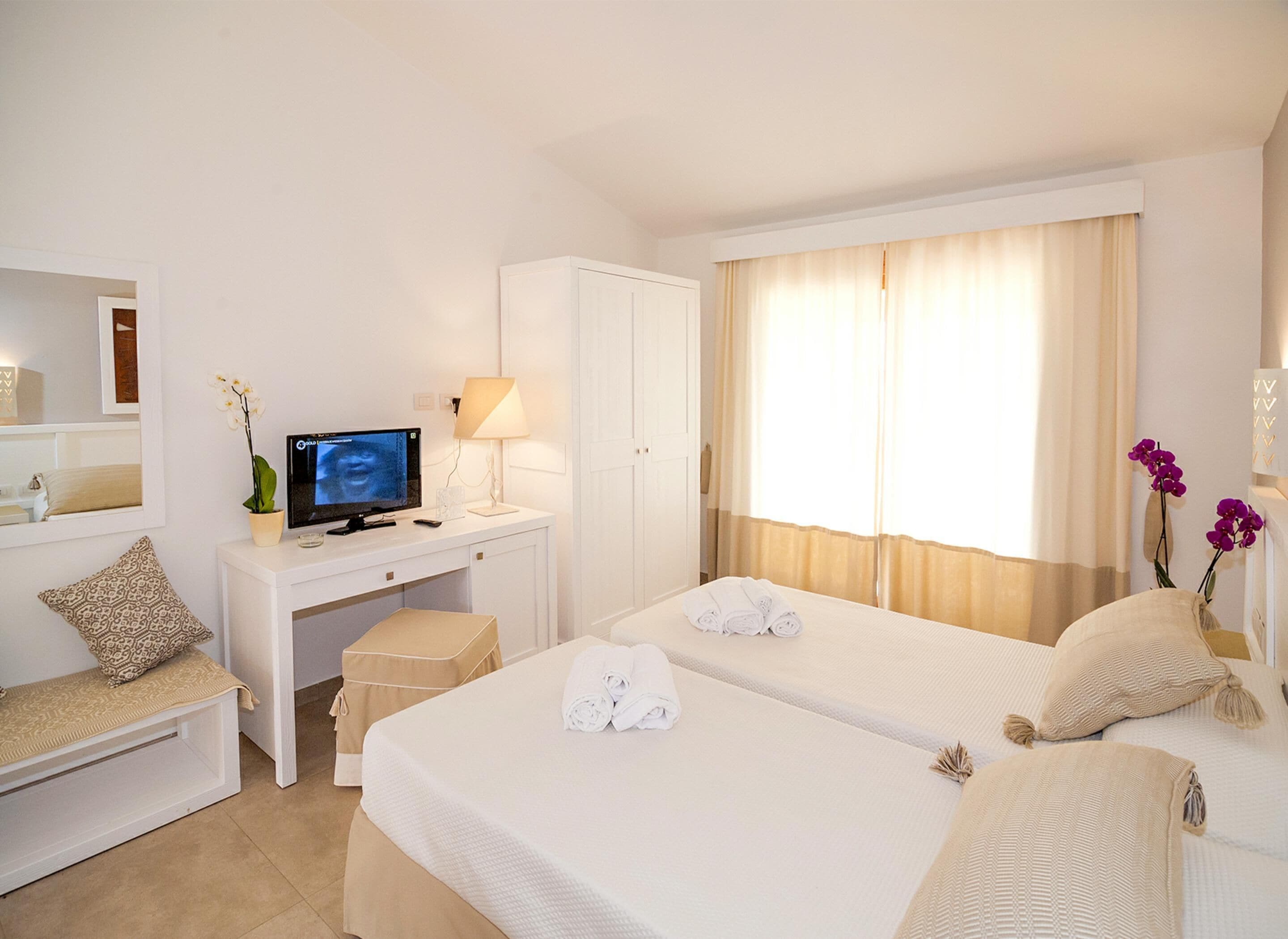 A hotel room with two beds, a desk with a computer, a chair, and a small table with a lamp.