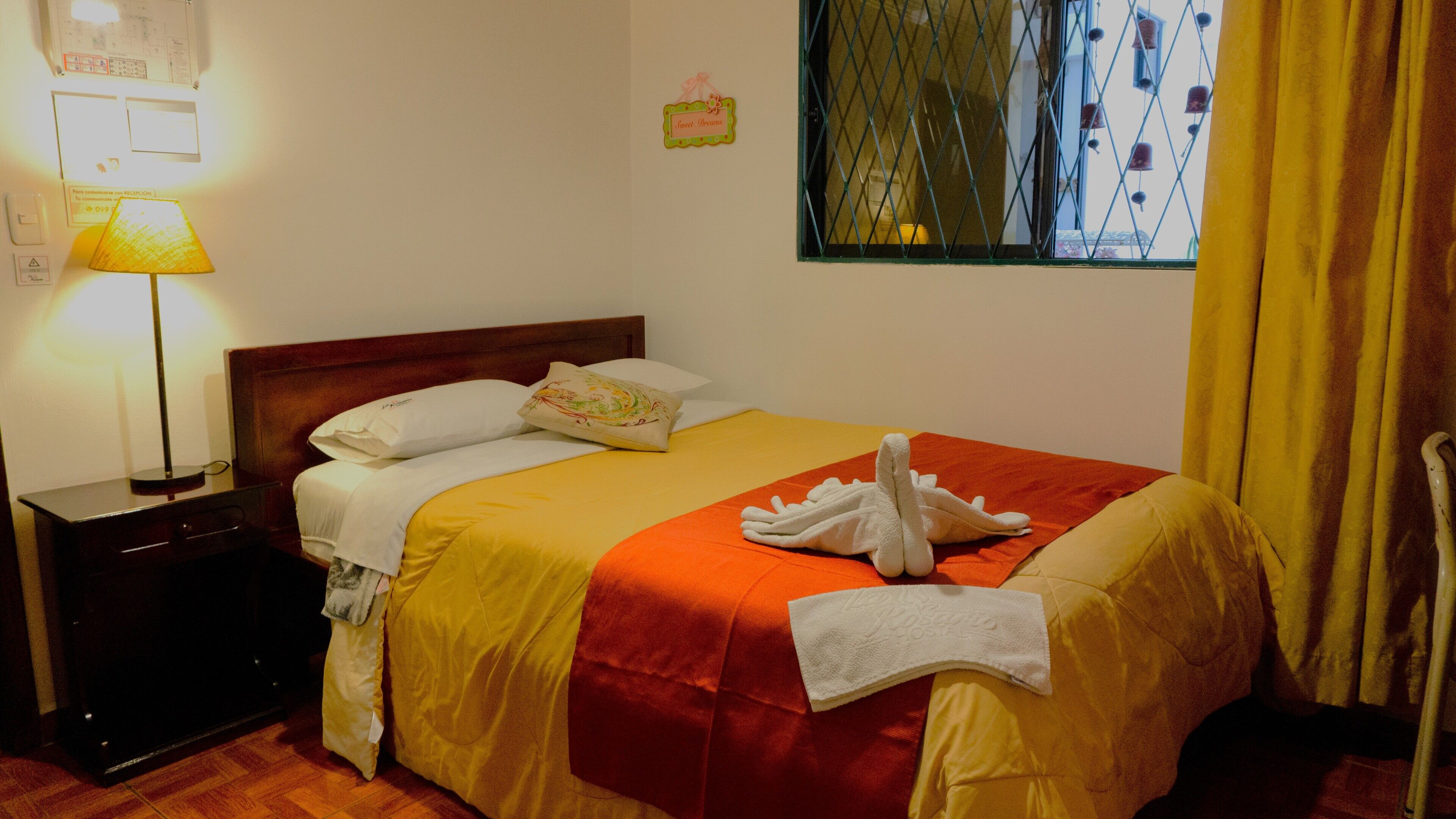 A neatly made bed with a red and yellow bedspread, a white towel, and a white bathrobe on the bed. There is a nightstand with a lamp and a wall clock.