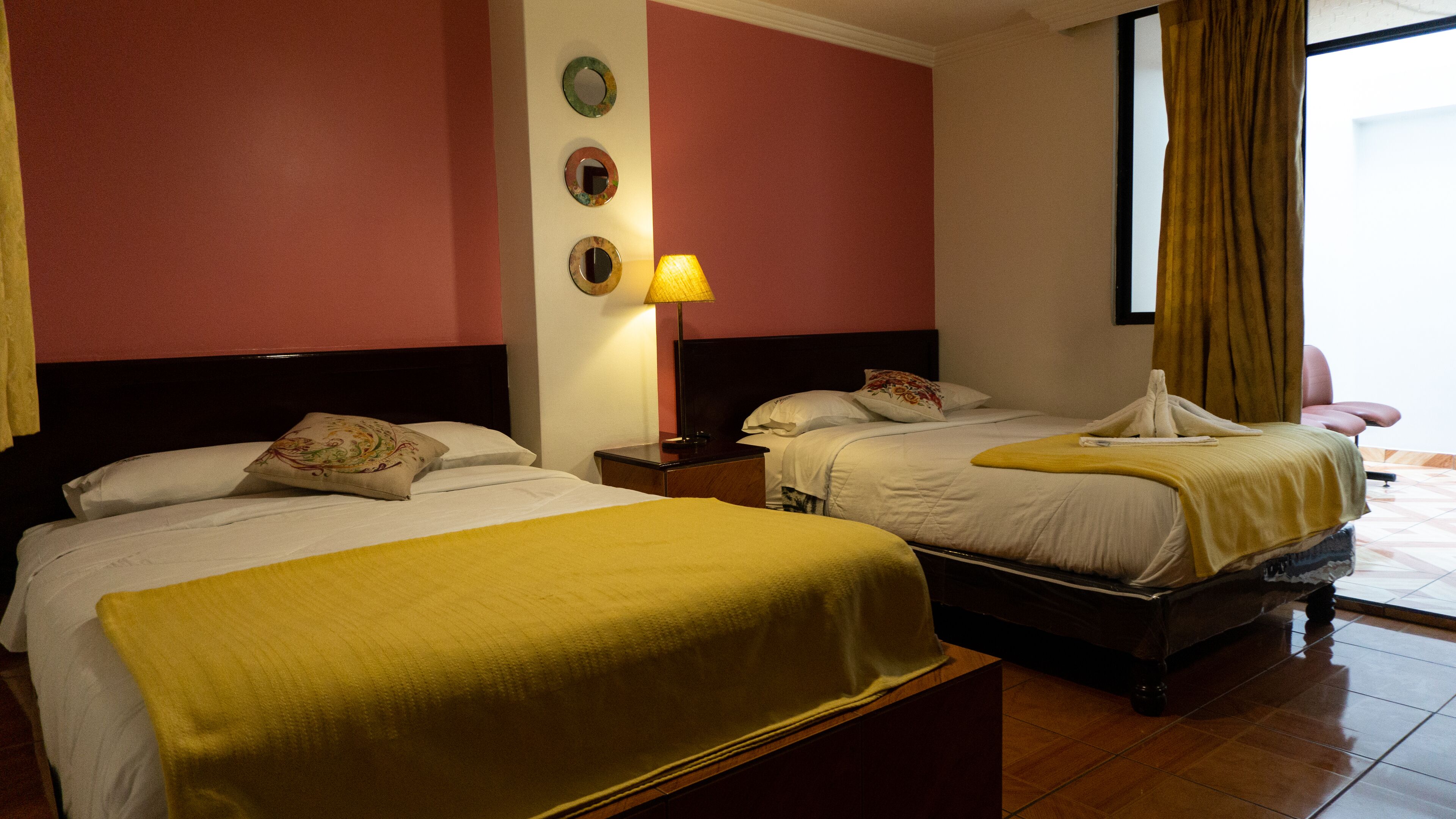 Quarto de hotel com duas camas, uma mesinha de cabeceira, uma luminária e uma janela com cortinas.