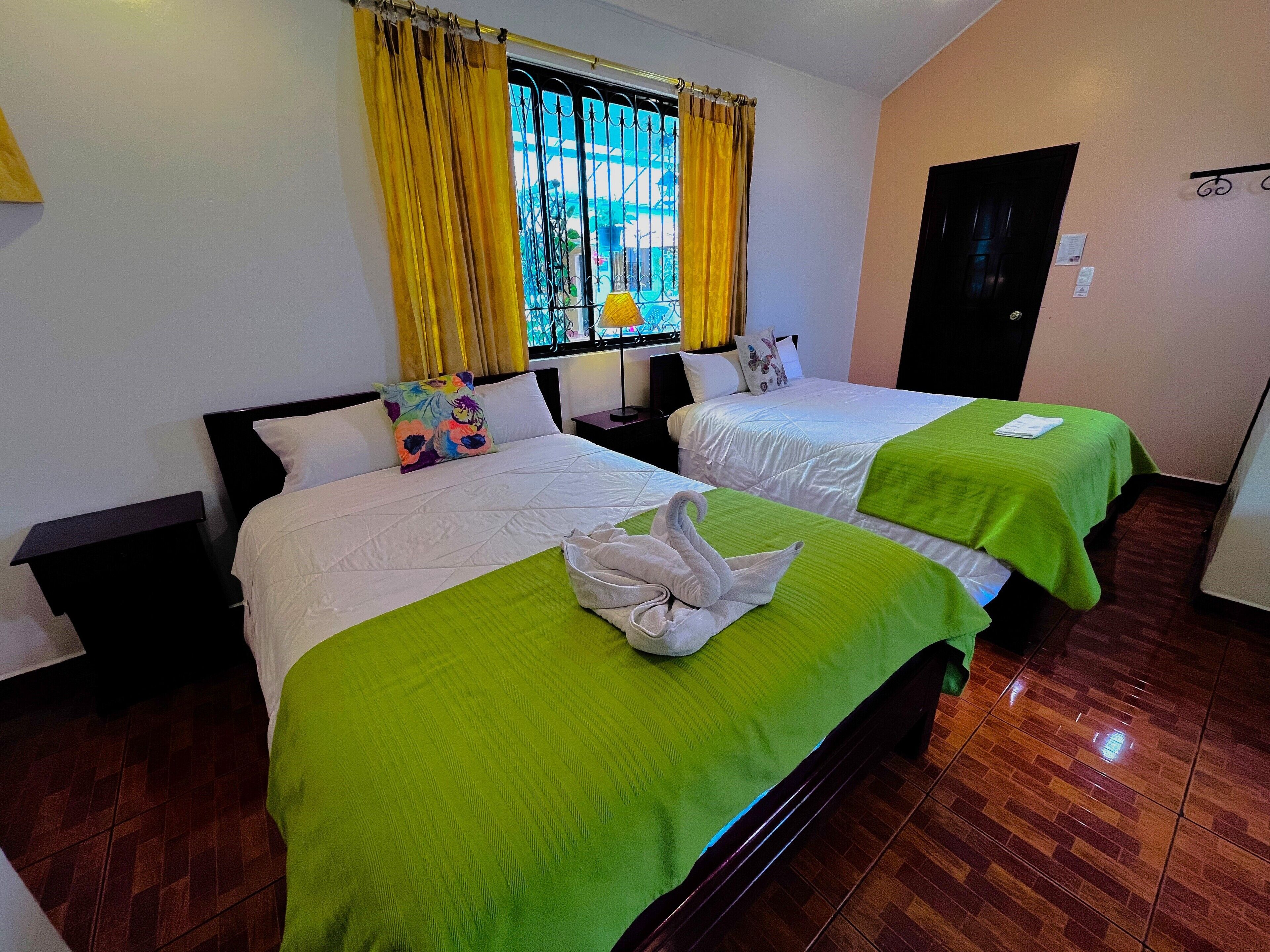 A hotel room with two beds, each with a green bedspread and white linens. There is a window with yellow curtains, a door, and a small table with a lamp.