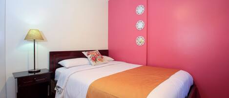 Una cama bien hecha con una colcha blanca y naranja, una lámpara de noche y una mesita de noche con un cajón.