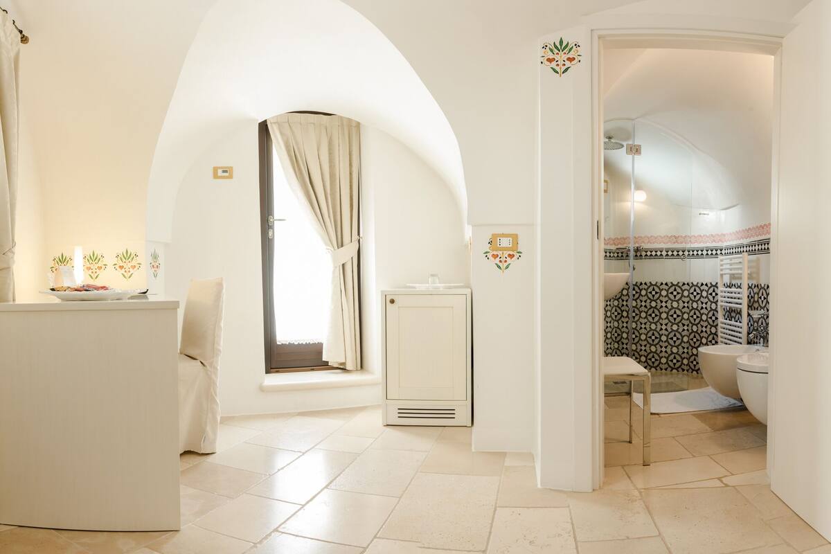 A hotel room with a bed, a small table with a vase, a window with curtains, a bathroom with a shower and toilet, and a kitchenette.