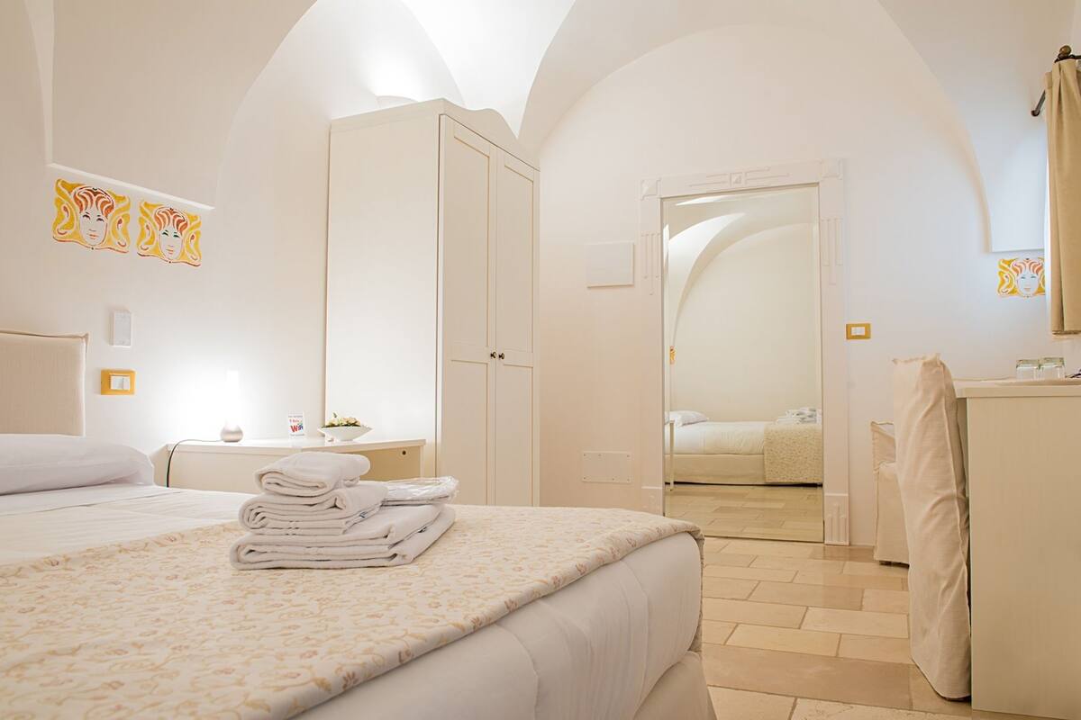 A hotel room with a bed, a bedside table with a lamp, a bathroom with a mirror, and a view of another room through an arched doorway.