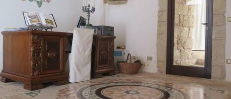 Una habitación con pared de piedra, un armario de madera, un paño blanco cubriendo una silla, un candelabro y un suelo con diseño.