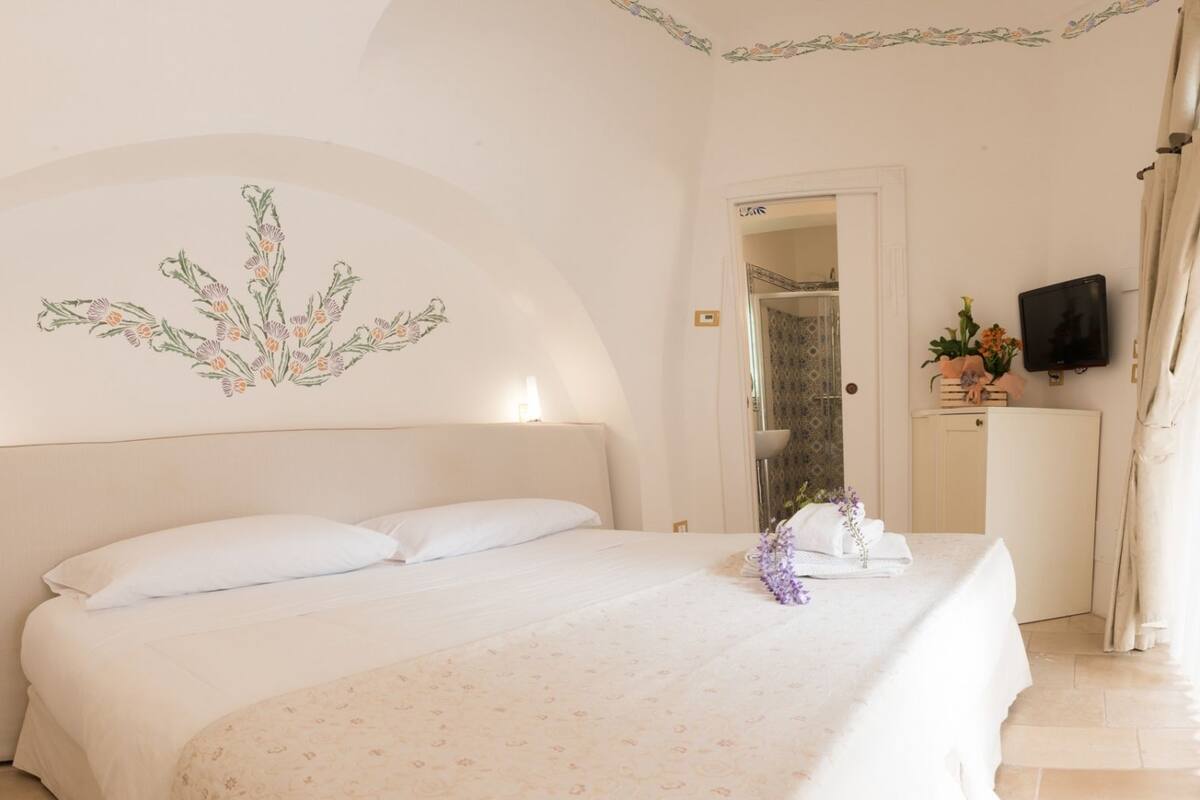 A bedroom with a large bed, a headboard, a wall-mounted TV, and a bathroom visible through an open door.