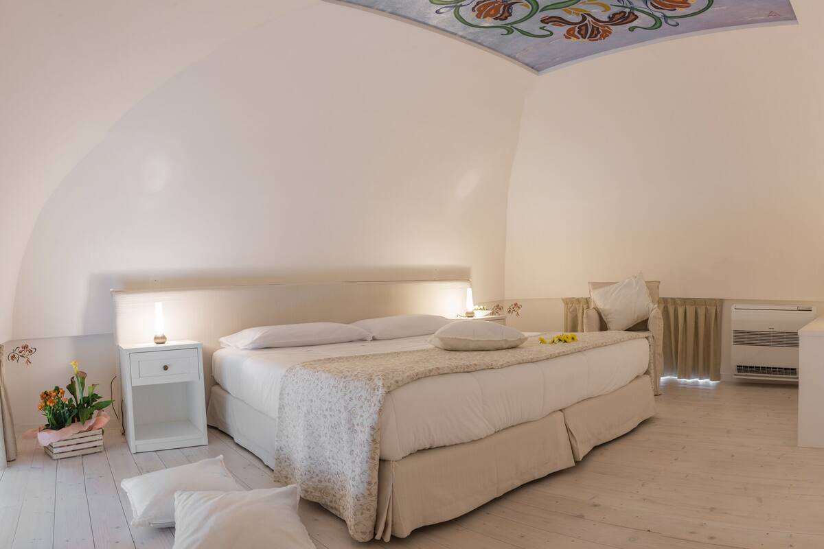 A spacious bedroom with a large bed, bedside tables, a desk, and a ceiling mural.