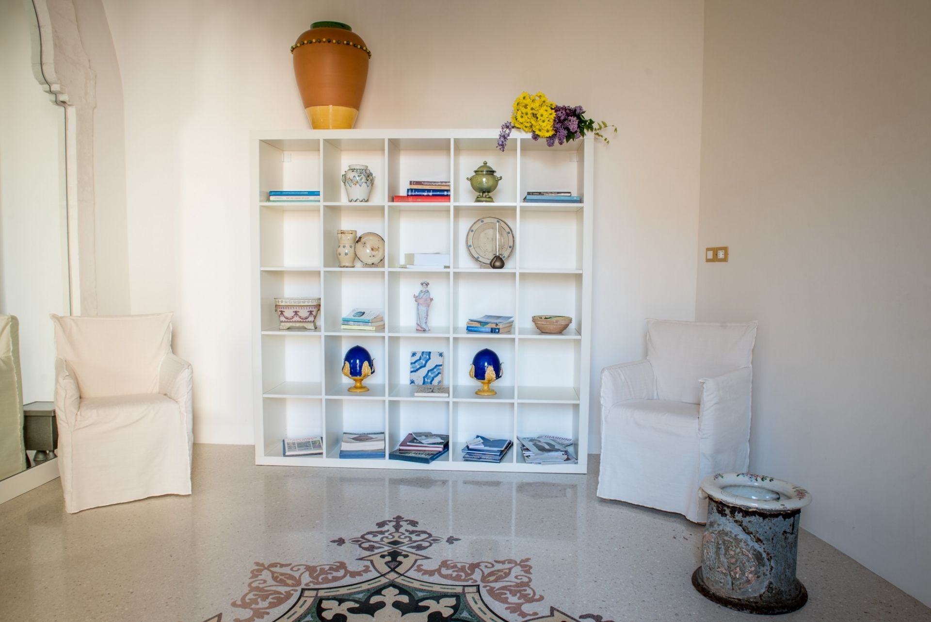 Un salon avec une bibliothèque blanche, deux fauteuils blancs, un tapis à motifs et des toilettes au style rustique.