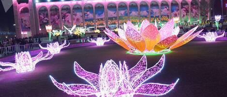 A large illuminated lotus flower installation in front of a building with arched windows and a mural.