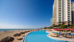 A resort with a large swimming pool, beach umbrellas, and a high-rise building.
