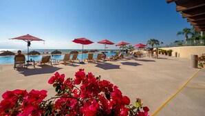 A poolside area with lounge chairs and red umbrellas, overlooking a clear blue sky and ocean.