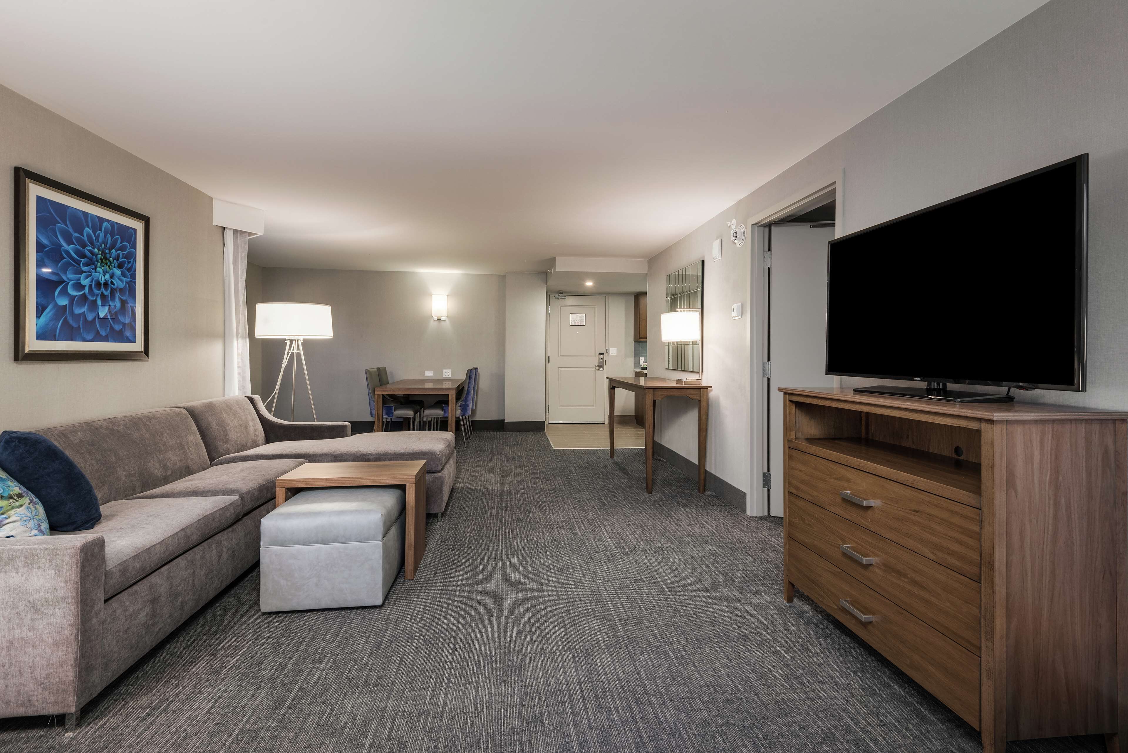A hotel room with a grey carpet, a grey sofa, a wooden dresser, a flat-screen TV, a desk with a lamp, and a door leading to a bathroom.