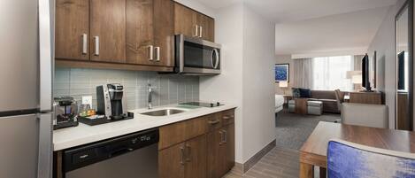 A modern kitchen with wooden cabinets, a dishwasher, a microwave, and a coffee maker. There is a dining area with a table and chairs, and a living room with a sofa and a TV.