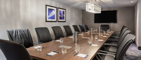 A conference room with a long wooden table, black leather chairs, and a large flat-screen TV on the wall.