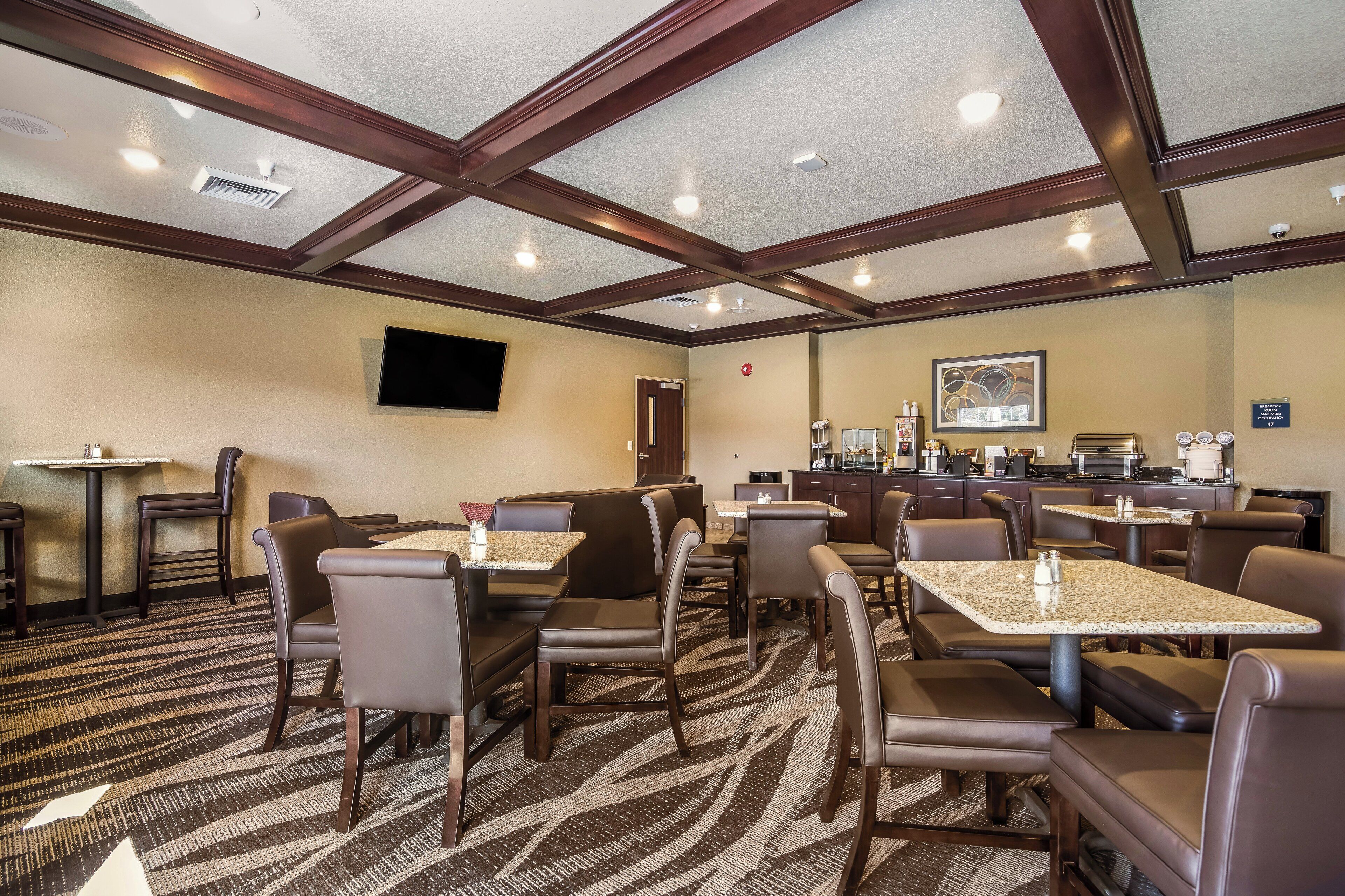 A hotel dining area with multiple tables and chairs, a kitchen in the background, and a television mounted on the wall.