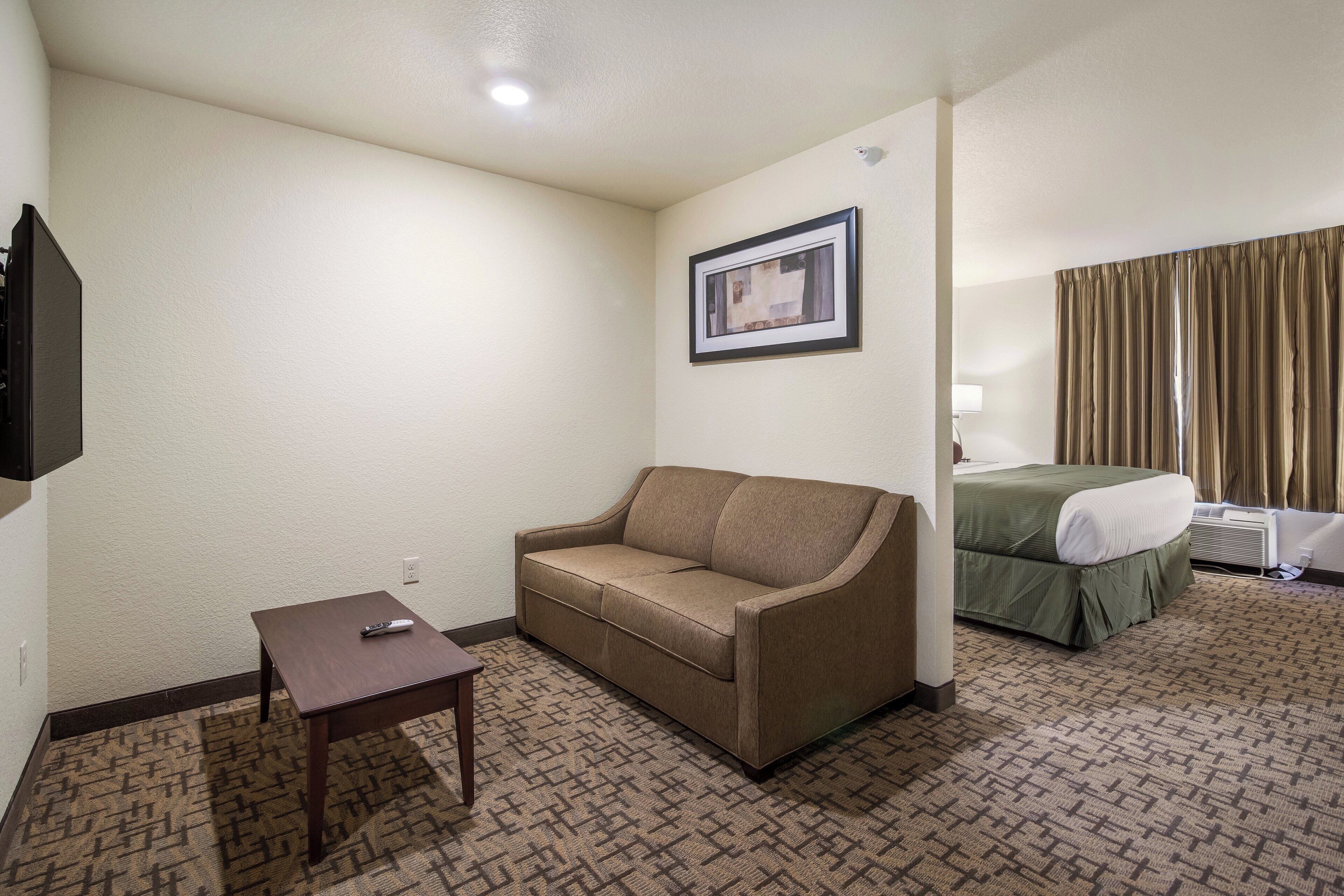 A hotel room with a sofa, a small table, a framed picture on the wall, and a bed with a green bedspread.