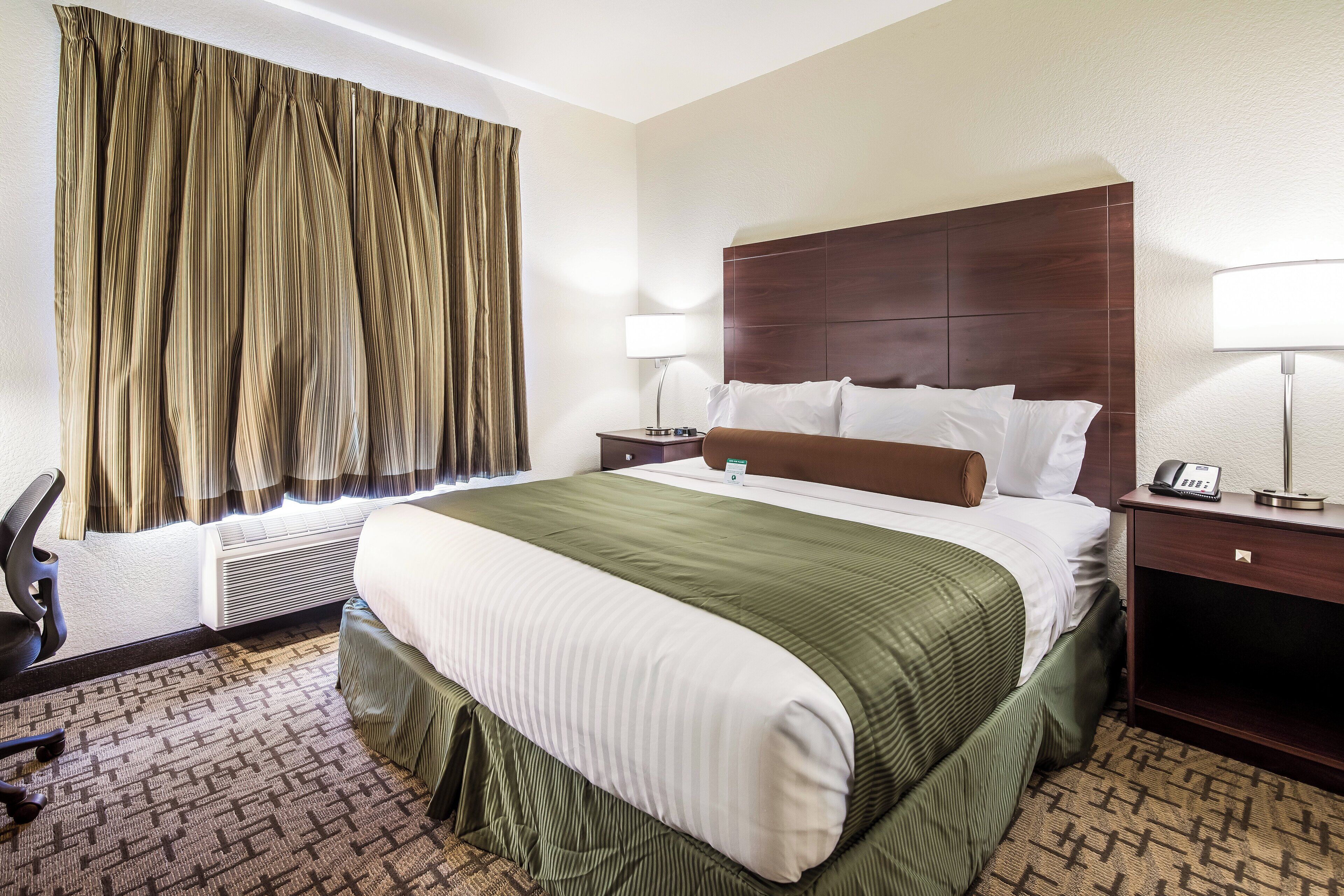 Una habitación de hotel con una cama grande, dos mesitas de noche, una silla y un ventanal con cortinas.