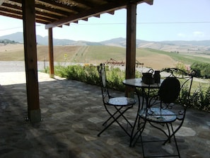 A covered outdoor seating area with stone flooring, metal chairs, and a table with a kettle and cup.