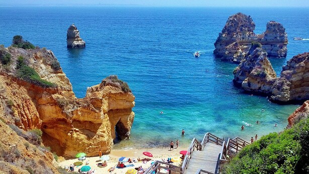 Une plage côtière bordée de falaises rocheuses, avec une eau d’un bleu limpide et un escalier en bois qui descend jusqu’au rivage.